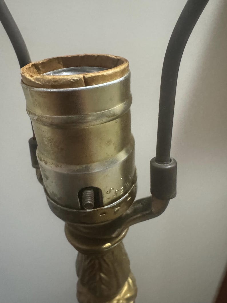 May include: A gold-colored lamp base with a decorative floral design. The lamp base has a standard light bulb socket with a cardboard insulator.