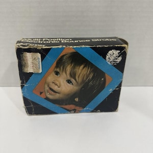 May include: A vintage JCPenney Multi-Position Electronic Bounce Strobe box. The box is black with a blue diamond-shaped frame containing a portrait of a smiling child. The box has some wear and tear.