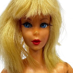 May include: A vintage doll with blonde hair and blue eyes. The doll has red lipstick and a fair complexion. The doll's hair is styled with a fringe. The doll is a collectible item.