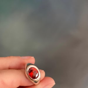 Może przedstawiać: Srebrny pierścionek z dużym, owalnym, ciemnoczerwonym kamieniem szlachetnym. Kamień jest osadzony w gładkiej, polerowanej srebrnej oprawie. Pierścionek jest trzymany na rozmytym tle.