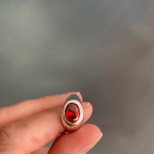 May include: A silver ring featuring a large, oval-cut, red gemstone. The gemstone is set in a smooth, polished silver band. The ring is held up against a blurred, neutral background.
