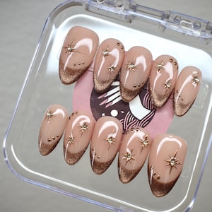 May include: A set of ten nude-colored false nails with gold glitter and a gold accent at the tip. The nails are arranged in a clear plastic case.