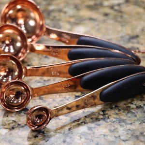 May include: Set of five copper measuring spoons with black rubber handles. The spoons are nested together and the handles are visible.
