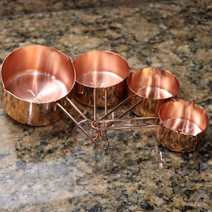 May include: A set of four copper measuring cups. The cups are linked together by a copper ring and handles. The copper has a shiny, reflective surface. The background is a speckled, brown countertop.
