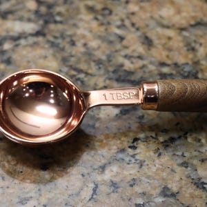 May include: A copper-colored measuring spoon with a wooden handle. The spoon is marked "1 TBSP" on the handle. The handle is a light brown color with a wood grain pattern. The spoon is on a speckled brown surface.