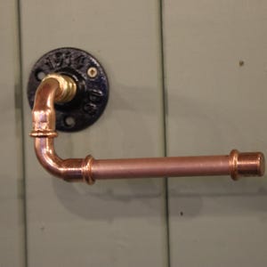 May include: Copper-colored industrial-style toilet paper holder. The holder is made of copper pipes and fittings, mounted on a black circular plate. The fixture is attached to a light green wooden surface.