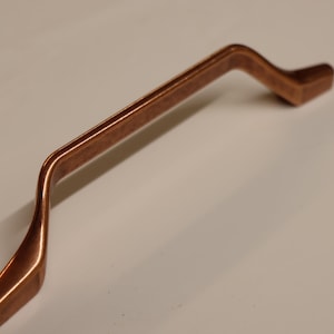 May include: A copper cabinet pull with a curved design. The pull is smooth and has a brushed finish.