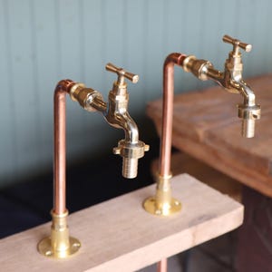 May include: Two brass spigots mounted on copper pipes and attached to a wooden surface. The spigots have a traditional design with a cross-shaped handle.