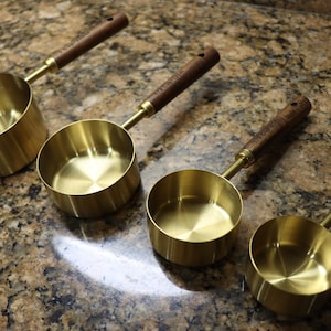 May include: Four brass measuring cups with wooden handles are arranged on a speckled countertop. The cups are different sizes, with a shiny, golden finish. The handles are a dark brown colour.