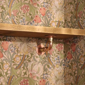 May include: A wooden shelf with a copper pipe bracket against a floral wallpaper background. The shelf is a light brown color and appears to be made of reclaimed wood. The copper pipe bracket is a curved design.
