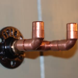 Könnte beinhalten: Ein Wandhaken aus Kupferrohr mit schwarzem Flansch. Der Haken besteht aus Kupferrohren und -fittings mit zwei nach oben gerichteten Armen. Der Flansch ist rund mit mehreren Löchern zur Montage. Die Vorrichtung ist zum Aufhängen von Gegenständen bestimmt.
