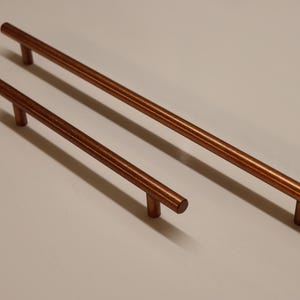 May include: Two copper cabinet pulls, one longer than the other, on a white surface.
