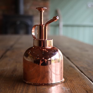 May include: A copper plant mister with a long, thin spout and a handle. The mister is sitting on a wooden table.