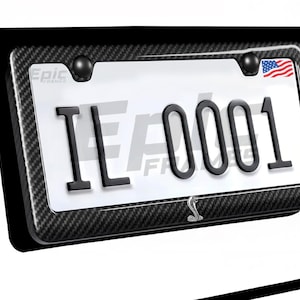 Könnte beinhalten: Ein Nummernschildrahmen mit Karbonfaser-Design. Das Kennzeichen lautet "IL 0001" in schwarzer Schrift. Eine amerikanische Flagge ist in der oberen rechten Ecke zu sehen. Der Rahmen trägt den Text "Epic Frames" und ein Cobra-Logo.