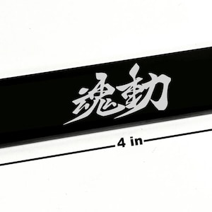 Puede incluir: Un objeto rectangular negro, de 10 cm de largo y 2,5 cm de ancho, con caracteres japoneses blancos. El objeto es brillante y parece ser un artículo decorativo o un componente de un producto más grande.