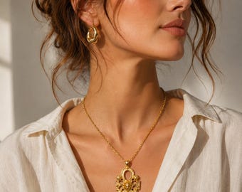 May include: A gold-toned necklace featuring an ornate ankh-shaped pendant with intricate embossed detailing and a central circular motif, suspended from a delicate gold chain, paired with matching small, smooth, rounded gold hoop earrings.