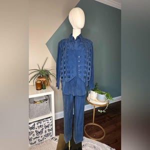 May include: A blue velvet three-piece suit displayed on a mannequin. The suit includes a long-sleeved button-down shirt, a vest with a square pattern, and matching pants. The mannequin is set against a neutral background with plants and a side table.