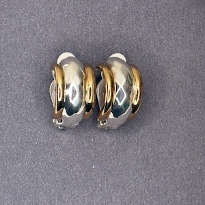 May include: A pair of clip-on earrings featuring a silver-tone base and gold-tone detailing. The earrings have a curved, geometric shape with a shiny finish. The gold-tone sections encircle the silver base, producing a layered appearance. The earrings are presented on a grey surface.