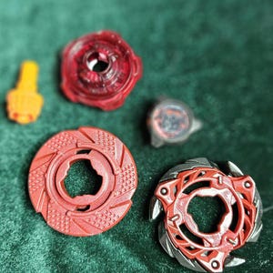 Puede incluir: Varios trompos y componentes de juguete rojos y negros est&aacute;n dispuestos sobre una superficie verde. Los trompos tienen varios dise&ntilde;os, incluyendo formas circulares y multicapa. Un componente es amarillo.