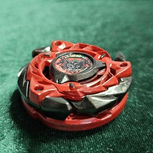 Puede incluir: Un juguete giratorio rojo y negro con un emblema de lobo en el centro. El juguete tiene un dise&ntilde;o multicapa con bordes afilados y una forma circular. La pieza central es negra con un gr&aacute;fico de lobo rojo.