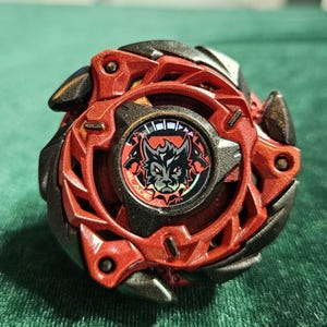 Puede incluir: Un juguete giratorio rojo y negro con un gr&aacute;fico de lobo en el centro. El juguete tiene un dise&ntilde;o multicapa con bordes afilados y una forma circular. El centro presenta la palabra "BLADE" y una ilustraci&oacute;n de lobo.