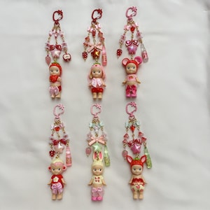 Pu&ograve; includere: Sei portachiavi con piccole figure di bambole, ognuna adornata con ciondoli decorativi nei toni del rosa, rosso e oro. Le bambole sono vestite con abiti a tema, tra cui fragole, coniglietti e un topo. Ogni portachiavi ha un moschettone.