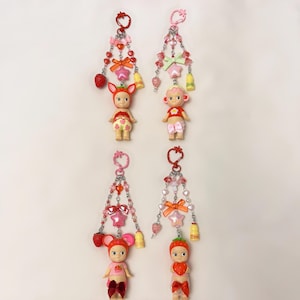 Puede incluir: Cuatro llaveros con peque&ntilde;as figuras de mu&ntilde;ecas, cada una adornada con cuentas, lazos y abalorios. Las mu&ntilde;ecas visten trajes con tem&aacute;tica de fresas, lazos y frutas. Los llaveros presentan detalles en rosa, rojo y plata, con abalorios en forma de coraz&oacute;n, estrella y campana.