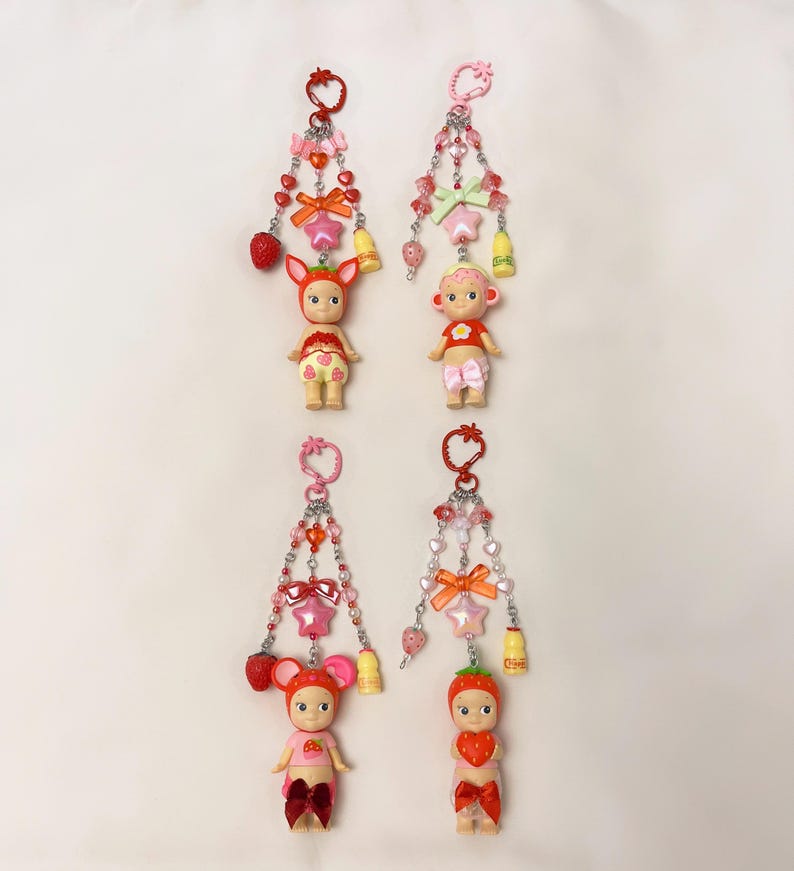 Puede incluir: Cuatro llaveros con peque&ntilde;as figuras de mu&ntilde;ecas, cada una adornada con cuentas, lazos y abalorios. Las mu&ntilde;ecas visten trajes con tem&aacute;tica de fresas, lazos y frutas. Los llaveros presentan detalles en rosa, rojo y plata, con abalorios en forma de coraz&oacute;n, estrella y campana.