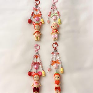 Puede incluir: Cuatro llaveros con peque&ntilde;as figuras de mu&ntilde;ecas, cada una adornada con dijes de fresas y estrellas, lazos y cuentas. Las mu&ntilde;ecas visten trajes con tem&aacute;tica de fresas y est&aacute;n suspendidas de un llavero rosa.