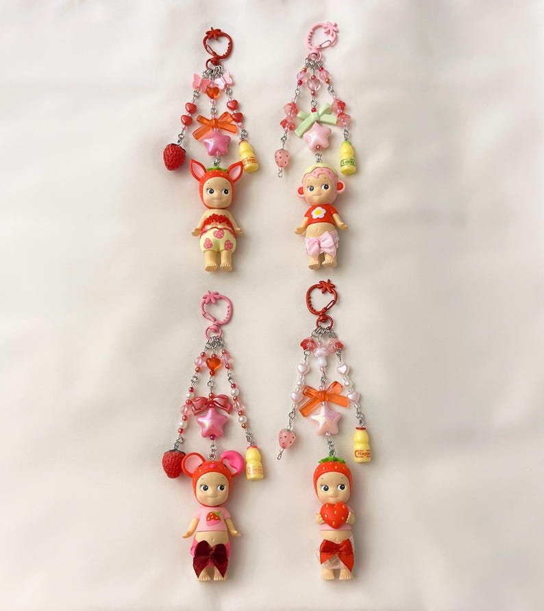 Puede incluir: Cuatro llaveros con peque&ntilde;as figuras de mu&ntilde;ecas, cada una adornada con dijes de fresas y estrellas, lazos y cuentas. Las mu&ntilde;ecas visten trajes con tem&aacute;tica de fresas y est&aacute;n suspendidas de un llavero rosa.