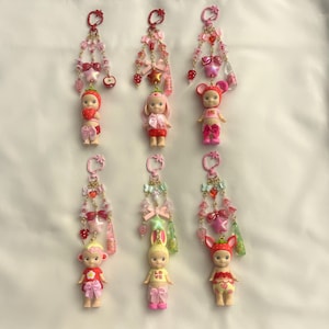 Pu&ograve; includere: Una collezione di portachiavi con piccole figure simili a bambole vestite con vari costumi a tema frutta e animali. Ogni portachiavi ha una catena di perline con fiocchi, ciondoli e un anello superiore. La tavolozza dei colori include rosa, rosso e giallo.