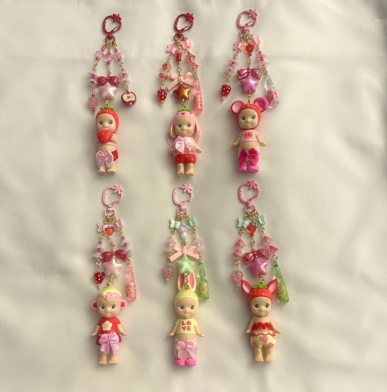 Pu&ograve; includere: Una collezione di portachiavi con piccole figure simili a bambole vestite con vari costumi a tema frutta e animali. Ogni portachiavi ha una catena di perline con fiocchi, ciondoli e un anello superiore. La tavolozza dei colori include rosa, rosso e giallo.