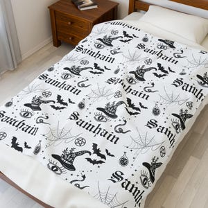 May include: White blanket with a repeating black pattern of witch hats, bats, spiderwebs, eyes, and the word "Samhain". The blanket is spread on a bed with a wooden frame and a nightstand in the background.
