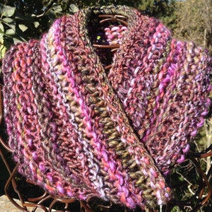 May include: A hand-knitted cowl scarf in shades of pink, purple, brown, and green. The chunky knit scarf is made of soft yarn and is designed to be worn around the neck. The scarf is displayed on a metal stand.