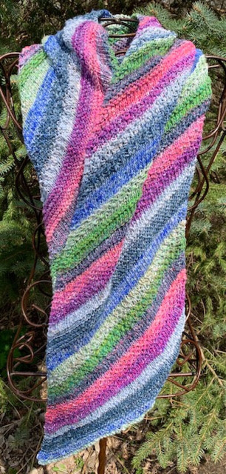 May include: A colourful striped knitted scarf in shades of pink, blue, green, and white. The scarf is folded and draped over a metal stand.