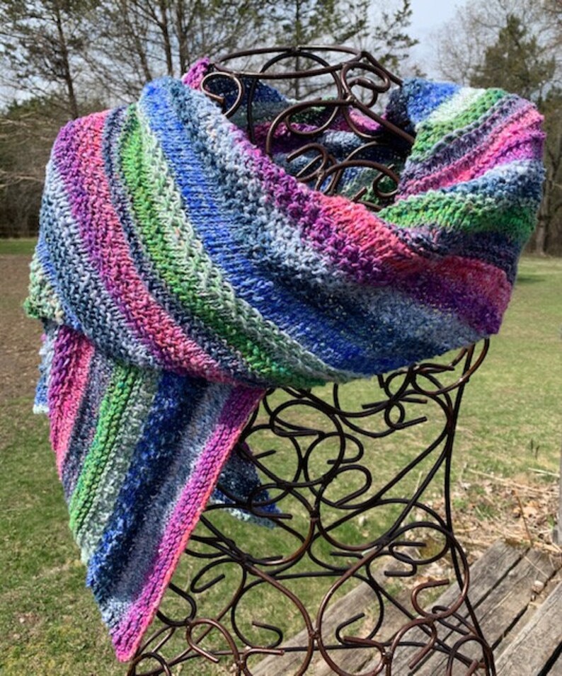 May include: A colourful striped knitted scarf in shades of blue, pink, green, and white. The scarf is draped over a decorative metal stand.