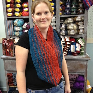 May include: A woman wearing a blue and orange knitted cowl scarf. The scarf is made with a textured stitch pattern. The woman is standing in front of a shelf filled with yarn.