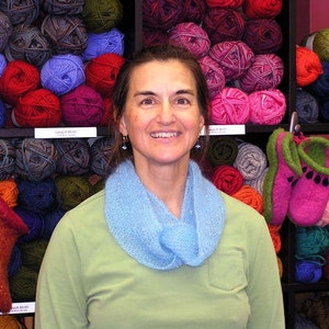May include: A woman wearing a light blue knitted scarf stands in front of shelves stocked with colorful yarn and knitted slippers. The shelves are labeled with the words "Lacy Knit Goods" and "Lacy Pride Yarns".