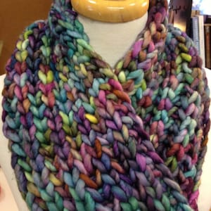 May include: A colorful knitted cowl with a variety of shades of blue, purple, green, and brown. The cowl is made with a chunky yarn and has a loose, drapey texture.