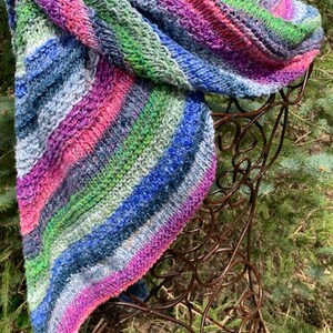 May include: A colourful striped knitted scarf in shades of pink, blue, green, and white. The scarf is draped over a brown metal stand.