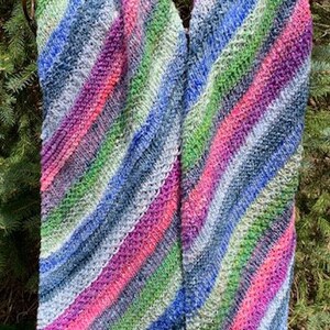 May include: A colourful striped knitted scarf in shades of blue, green, pink, and white. The scarf is draped over a metal stand and is made with a soft, textured yarn.