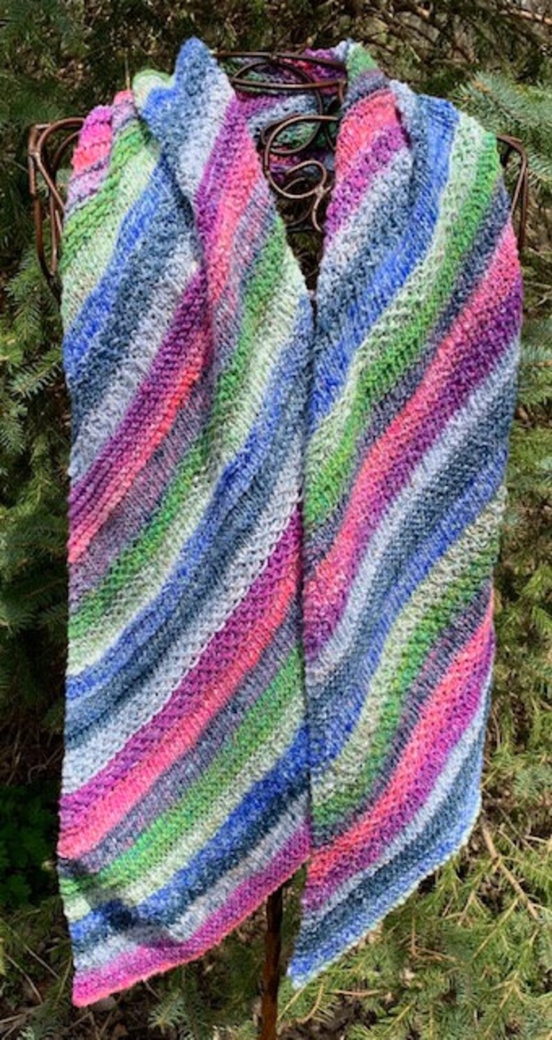 May include: A colourful striped knitted scarf in shades of blue, green, pink, and white. The scarf is draped over a metal stand and is made with a soft, textured yarn.