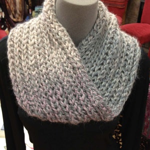 May include: A gray, pink, and white knitted cowl neck scarf. The scarf is displayed on a black mannequin.