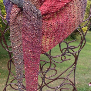 May include: A delicate, hand-knitted shawl in shades of red, blue, and brown with a beaded design. The shawl is draped over a decorative metal stand.