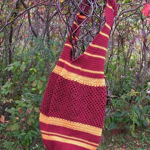 May include: A crocheted tote bag with maroon and orange stripes. The bag is hanging on a metal stand in front of a leafy background.