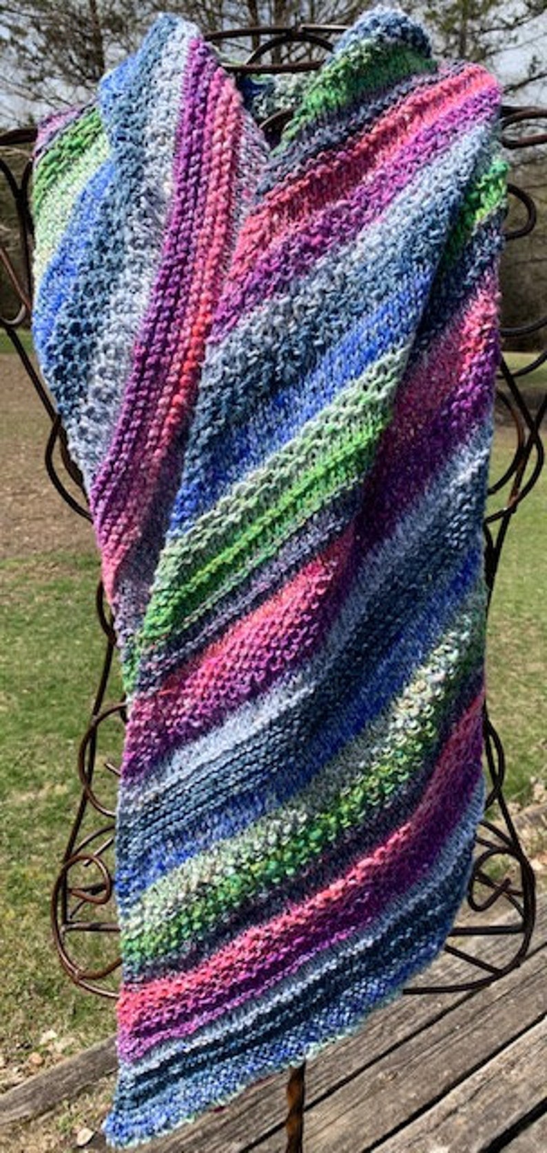 May include: A colourful striped knitted scarf with shades of blue, green, pink, and purple. The scarf is draped over a metal stand.