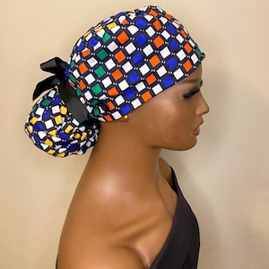 May include: A colorful surgical scrub hat with a checkered pattern in blue, green, orange, and yellow squares on a black background. The hat has a ponytail opening and a black ribbon tie.
