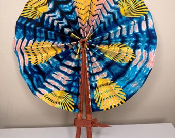 May include: A circular hand fan featuring a vibrant blue, pink, and white tie-dye pattern with repeating yellow shell-like motifs. The fan is attached to two brown leather-wrapped handles secured together with a brown leather strap at the base.
