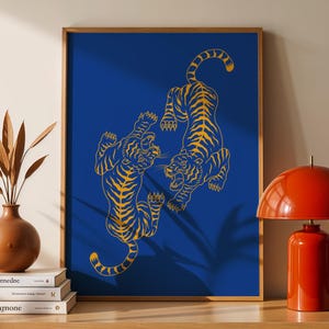 May include: A framed art print featuring two stylized gold tigers on a deep blue background. The tigers are depicted in a dynamic pose, facing each other. The artwork is displayed on a wooden cabinet with a red lamp.
