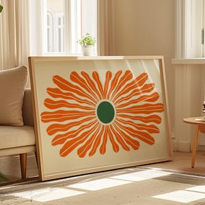 May include: A framed art print depicting a large, stylised orange flower with a green centre. The artwork is displayed on a light-coloured background and framed in a natural wood frame. The print is leaning against a wall in a well-lit room.